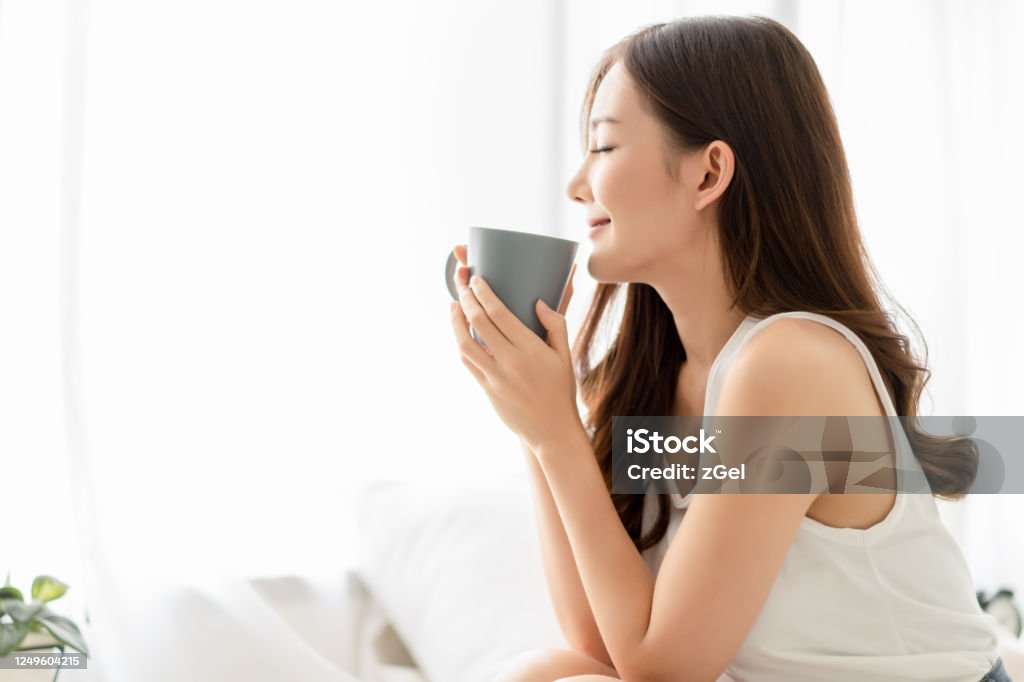 Happy smiling beautiful Asian woman enjoying first morning and smelling aroma of cup of coffee tea in her bedroom.Enjoying free and Relax time, Lazy day off concept. Drinking Stock Photo