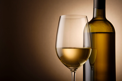 Unlabelled bottle and glass of white wine side by side highlighted over a shadowy brown background with copy space in a cropped close up