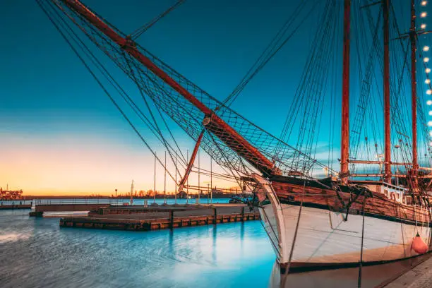 Photo of Helsinki, Finland. Old Wooden Sailing Vessel Ship Schooner Is Moored To City Pier, Jetty. Lighting At Evening Or Night Illumination