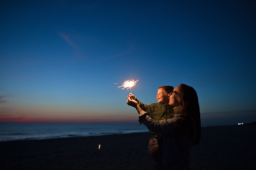 Happy little boy looking at sparkler with his parent near the sea