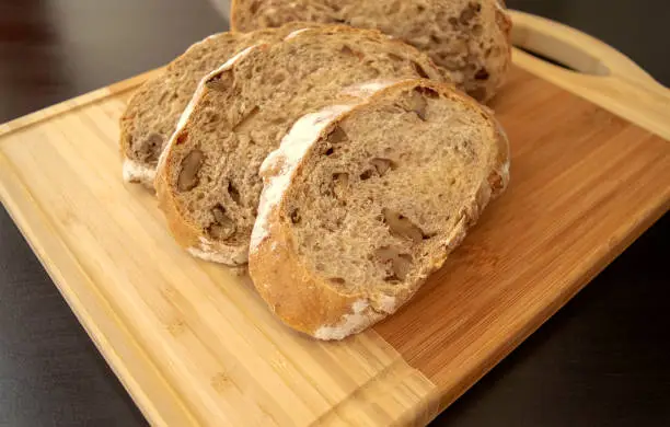 Photo of Sliced walnuts bread on wooden cutting board on dark brown background.