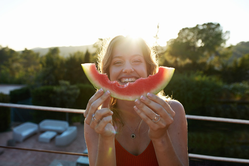 Front view with lens flare of woman in mid 20s smiling as she holds a partially eaten slice of watermelon on second story terrace of home in late afternoon.