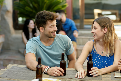 Male and female friends in mid 20s sitting side by side at outdoor table drinking beers and talking at weekend party.