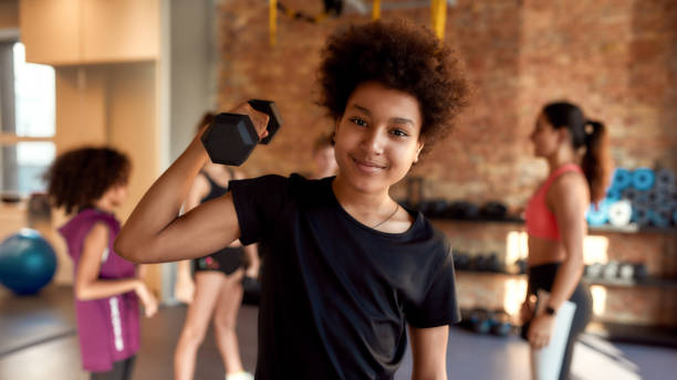 istock African american boy smiling at camera while exercising using dumbbell in gym together with female trainer and other kids. Sport, healthy lifestyle, physical education concept 1249645037