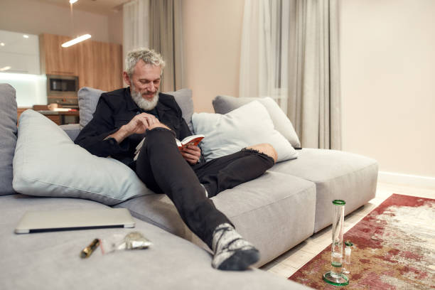 istock Time to read. Bearded middle-aged man reading a book while sitting on the couch. Bong or glass water pipe, buds in plastic bag and lighter near him. Cannabis and weed legalization concept 1249649311