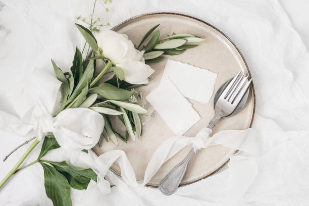 istock Festive summer wedding scene. Marble table setting with cutlery, olive branches, white peony flowers, stoneware plate and silk ribbon. Blank place cards mockups. Restaurant menu concept. Flat lay, top 1249652288