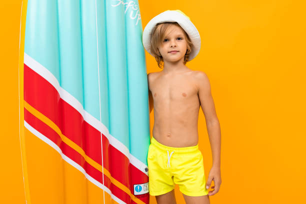 여름 파나마와 오렌지 배경에 팽창식 수영 보드가있는 노란색 수영 트렁크의 매력적인 유럽 금발 소년 - swimming trunks 뉴스 사진 이미지
