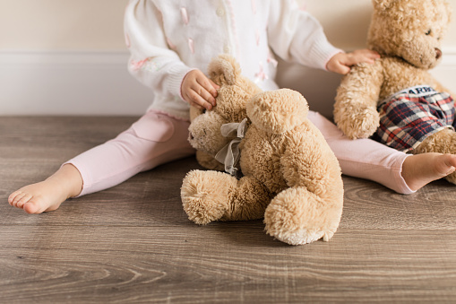 A 14-month-old baby girl, sitting on a wooden floor playing with her teddy bears. Toes, feet, and hands featured.