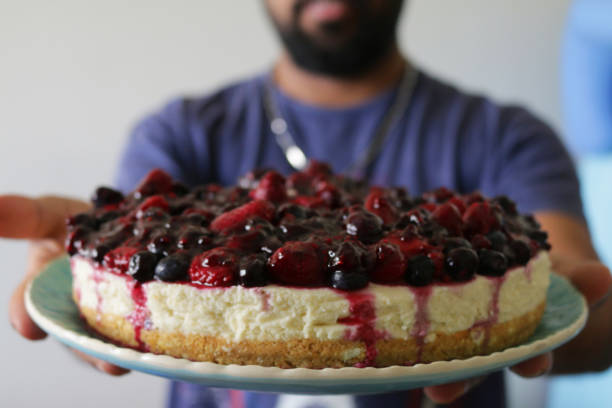 istock Image of handsome Indian man holding blackcurrant cheesecake with buttery biscuit base on patterned blue plate, vanilla mascarpone cream cheese dessert pudding topped with cherry jam coulis and fresh blackcurrants 1249697192