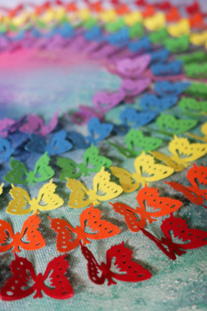 Stock photo of gay themed homemade crafting art work, using rainbow butterfly punch with cut out card and paper butterflies, embossed and glued on abstract watercolour painting that has been painted on a stretched canvas in circle shape. This original craft art work of rainbow coloured made of multicoloured cut out butterfly shapes (Monarch paper butterflies - purple, blue, green, orange, yellow and red colours) is like the rainbow LGBTQI gay flag, with the colourful arts and crafts project displayed on wall as background wallpaper. imagen de la obra de arte artesanal casero de temática gay, utilizando el ponche de mariposa arco iris con tarjeta cortada y mariposas de papel, en relieve y pegado en pintura de acuarela abstracta pintada sobre un lienzo estirado en forma de círculo. ar