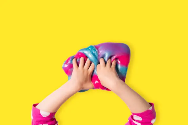 Child hands and colorful pink, blue and purple shiny slime. Child girl plays with slime isolated on bright yellow background. Home educational games concept. Top view, flat lay banner.