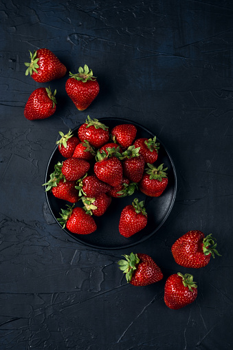 Ripe red strawberries in a black plate on a dark blue background. Sweet dessert from fresh berries on the table. Top view.