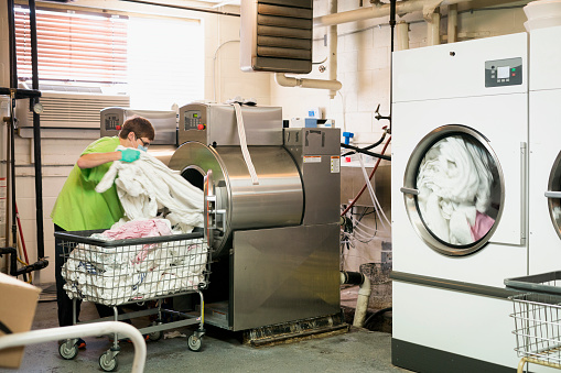 A red-haired female housekeeper wearing a face mask and green shirt removes sheets from a giant washing machine in the laundry room during the Corona virus pandemic, Midwest, USA