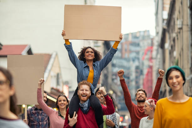 Happy group of people holding blank banner during strike Group of multiethnic people on city street holding blank cardboard placard celebrating victory during a protest. Young group of content men and smiling women marching through a city. People protesting on road with empty sign. protest stock pictures, royalty-free photos & images