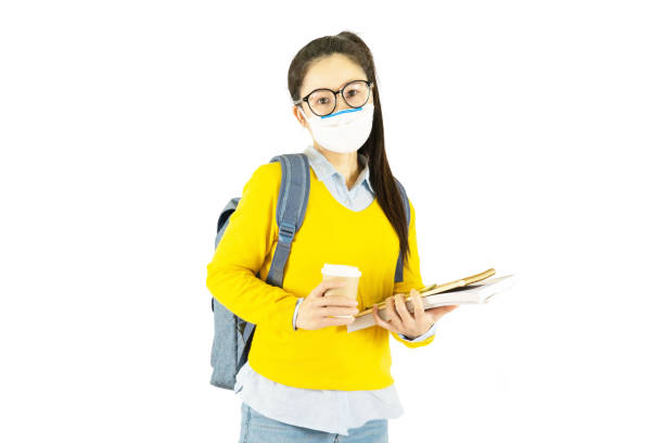 医療フェイスマスクを着用した眼鏡をかけた若いアジアの学生女性、白い背景に隔離されたウイルスの流行の下で学校に行くためにバッグとラップトップを運ぶ - collegian ストックフォトと画像