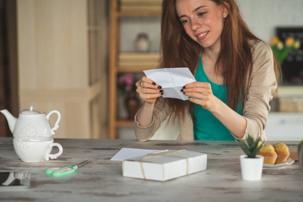 Young woman reading a letter she got Copy space shot of a happy young woman sitting at the table in her home and reading a letter she got with a gift box from a friend during quarantine. gift tag note stock pictures, royalty-free photos & images