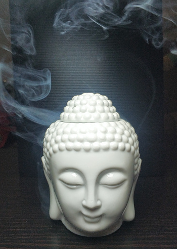 A close up shot of a ceramic sculpture based on buddha face with eyes closed, placed on a dark brown wooden table with light smoke effects in the background.