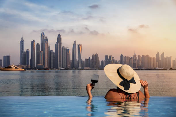 A woman with a hat stands at the edge of a pool and enjoys the view to the skyline of the Dubai Marina A elegant woman with a hat and a drink in her hand stands at the edge of a infinity pool and enjoys the view to the skyline of the Dubai Marina during sunset, UAE dubai photos stock pictures, royalty-free photos & images