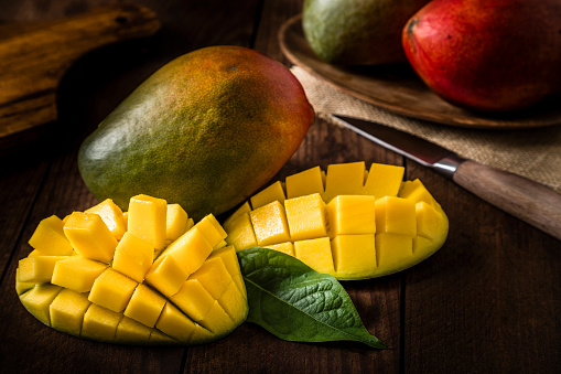 Closeup of one whole and one sliced ​​mango on a dark brown wooden table. The  objects are at the lower left corner and at the background are two more mangoes on a wooden tray, a kitchen knife and a wooden cutting board. Predominant colors are yellow, brown and green. Low-profile DSLR photo taken with Canon EOS 6D Mark II and Canon EF 24-105mm f / 4L