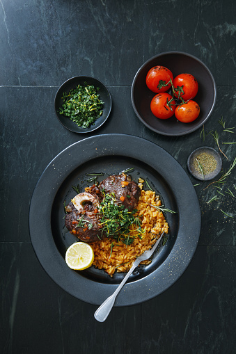 Traditional Italian Osso Buco with Creamy Saffron Risotto dish topped with fresh parsley, garlic and lemon zest. Flat lay top-down composition on dark background.