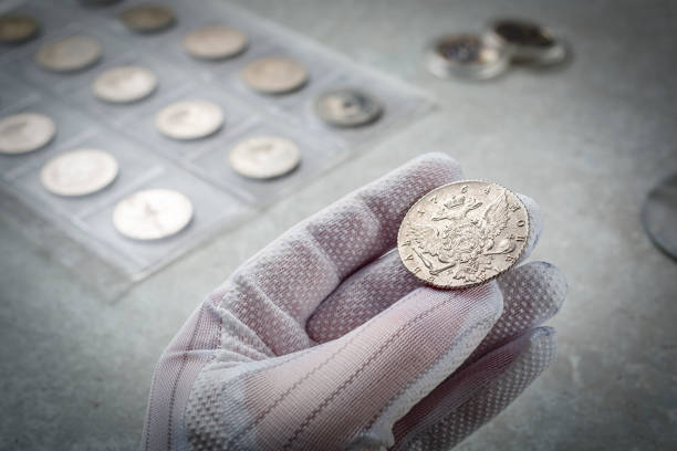 Numismatics. Old collectible coins made of silver on a wooden table.  A collector in special gloves holds an old coin. Numismatics. Old collectible coins made of silver on a wooden table.  A collector in special gloves holds an old coin. coin collection stock pictures, royalty-free photos & images