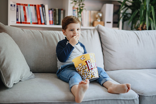Toddler boy watching TV and eating popcorns at home