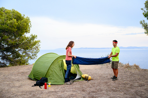 Smiling camping friends unfolding sleeping bag and setting up tent on cliff overlooking Mediterranean Sea.