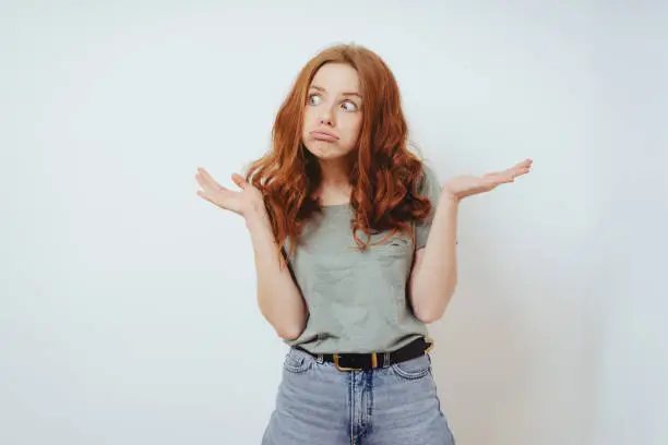 Confused young woman showing her bewilderment shrugging her shoulders and gesturing with her hands while pulling a face in front of a white wall with copy space