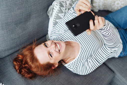 Cute vivacious young woman lying on a sofa on her back looking up at the camera with a mischievous grin while holding her mobile