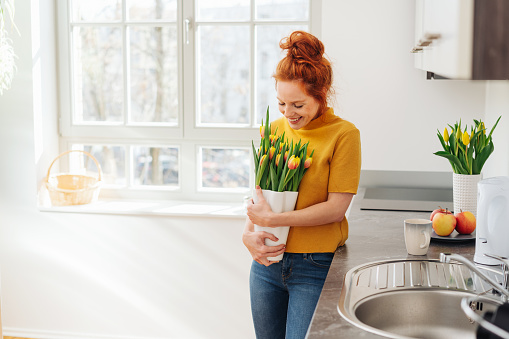 Smiling young lady with vase of flowers in her hands, standing and looking down at tulip bouquet, in kitchen interior with copy space