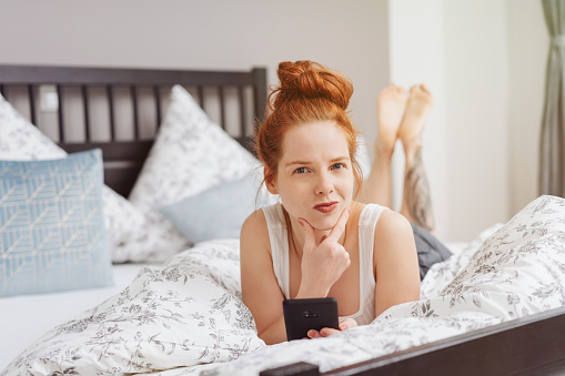 Thoughtful sceptical young redhead woman looking at the camera in disbelief as she relaxes on a bed at home with her mobile