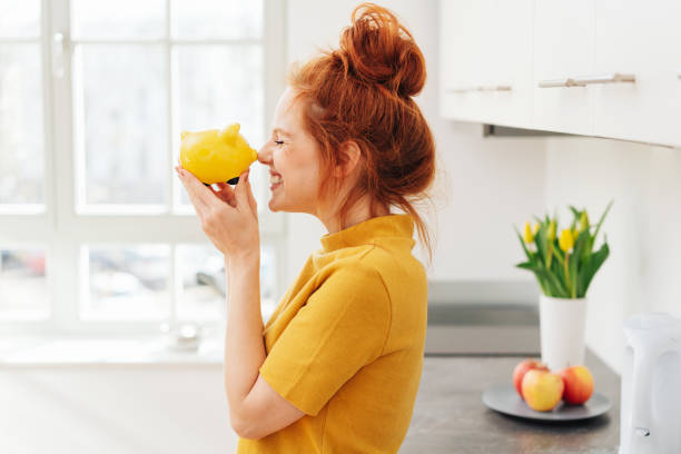 Smiling woman playing with piggy bank Smiling red-haired woman playing with yellow piggy bank in her hands, viewed from the side in bright room interior. Money savings concept savings stock pictures, royalty-free photos & images