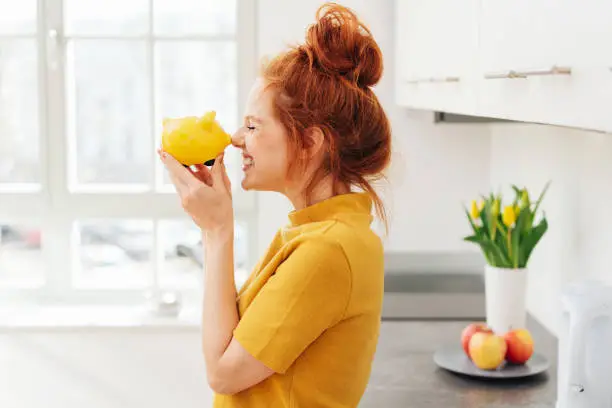 Smiling red-haired woman playing with yellow piggy bank in her hands, viewed from the side in bright room interior. Money savings concept