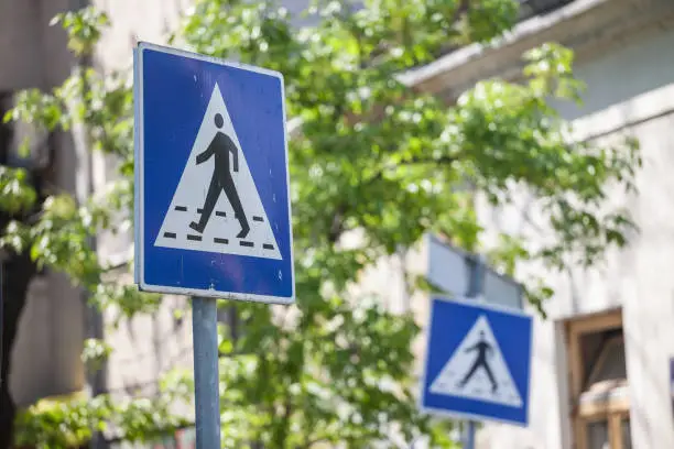 Picture of two roadsigns indicating the presence of a pedestrian crossing nearby, aimed at drivers, in a city environment, abiding by European standards.