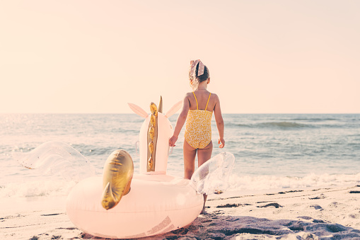 Beach scene - five years old little girl, wearing swimsuit and sunglasses, holding a giant pink inflatable unicorn. Sea and clear blue sky on the background.