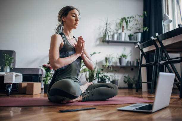 Beautiful woman meditating with online guru One beautiful young woman sitting in lotus pose on exercise mat in her living room and meditating. yoga lotus position meditating women stock pictures, royalty-free photos & images