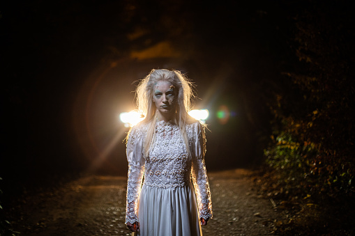 Zombie in wedding dress, with white hair, standing on a road at night with car behind her