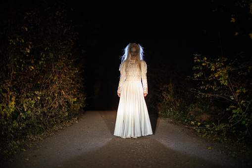 Witch in wedding dress, with white hair, standing on a road at night