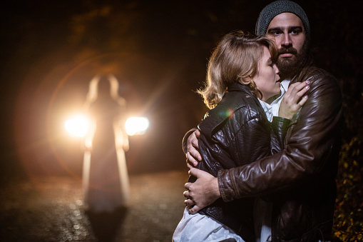 Man embracing his terrified girlfriend while ghost standing behind them, on road at night