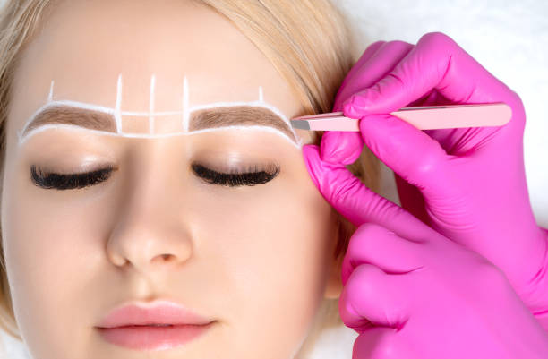 istock Woman Having Permanent Make-up Tattoo on her Eyebrows. Eyelash maker plucks eyebrows with tweezers. Professional makeup and cosmetology skin care. 1249779045