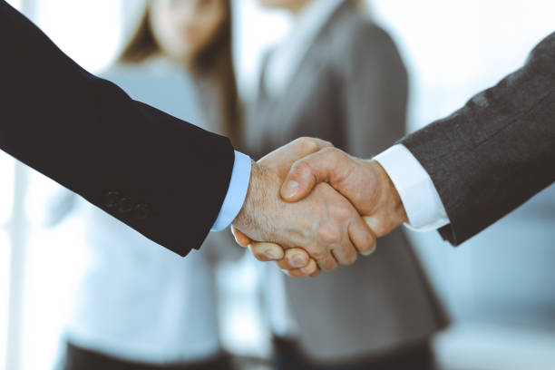 istock Business people shaking hands while standing with colleagues after meeting or negotiation, close-up. Group of unknown businessmen and women in modern office. Teamwork, partnership and handshake concept, toned picture 1249779252