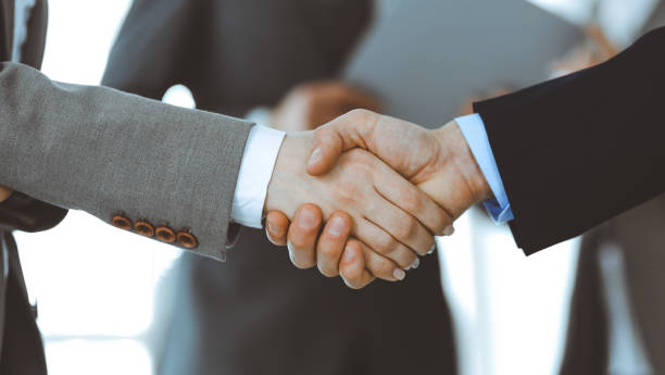 istock Business people shaking hands while standing with colleagues after meeting or negotiation, close-up. Group of unknown businessmen and women in modern office. Teamwork, partnership and handshake concept, toned picture 1249779264