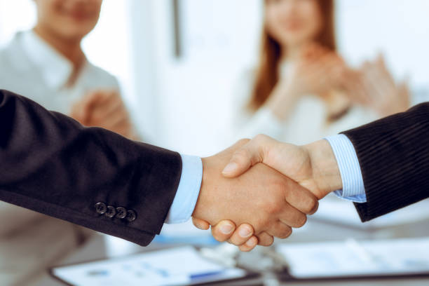 istock Business people shaking hands at meeting while theirs colleagues clapping and applauding. Group of unknown businessmen and women in modern white office. Success teamwork, partnership and handshake concept 1249779331