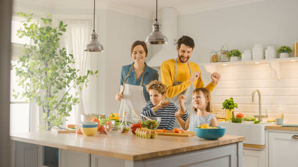 キッチンで:4人の家族が一緒に健康的な夕食を調理し、周りに愚か者と踊る。母、父、小さな男の子と女の子、サラダを準備し、野菜を切断します。かわいい子供たちが彼らの思いやりのあ� - child beauty mother little boys ストックフォトと画像