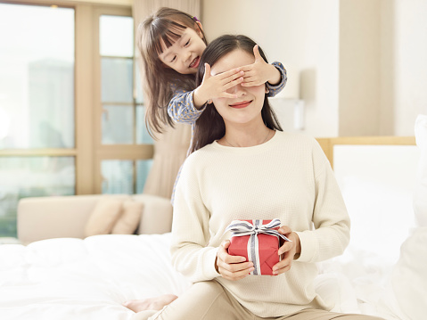 happy little asian girl giving mother a gift and covering mother's eyes with hands