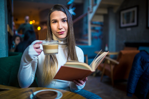 Young caucasian woman drinking coffee and reading a book in cafe
