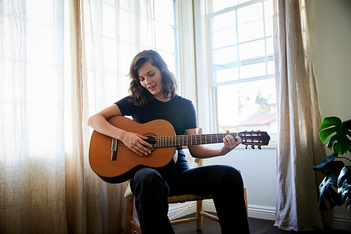 Young woman playing an acoustic guitar while sitting in a chair by a window at home