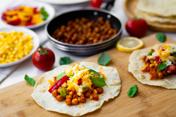 tortilla tacos with chickpeas, bell peppers, corn, tomatoes, basil leaves, green chili topped with cheddar and monterrey jack. on the background are ingredients, lemons, chickpeas in a small pan - mexico chili pepper bell pepper pepper imagens e fotografias de stock