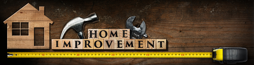 Text Home Improvement, made of wooden blocks on a wooden table with work tools (hammer, adjustable wrench and yellow tape measure) and stylized model house.