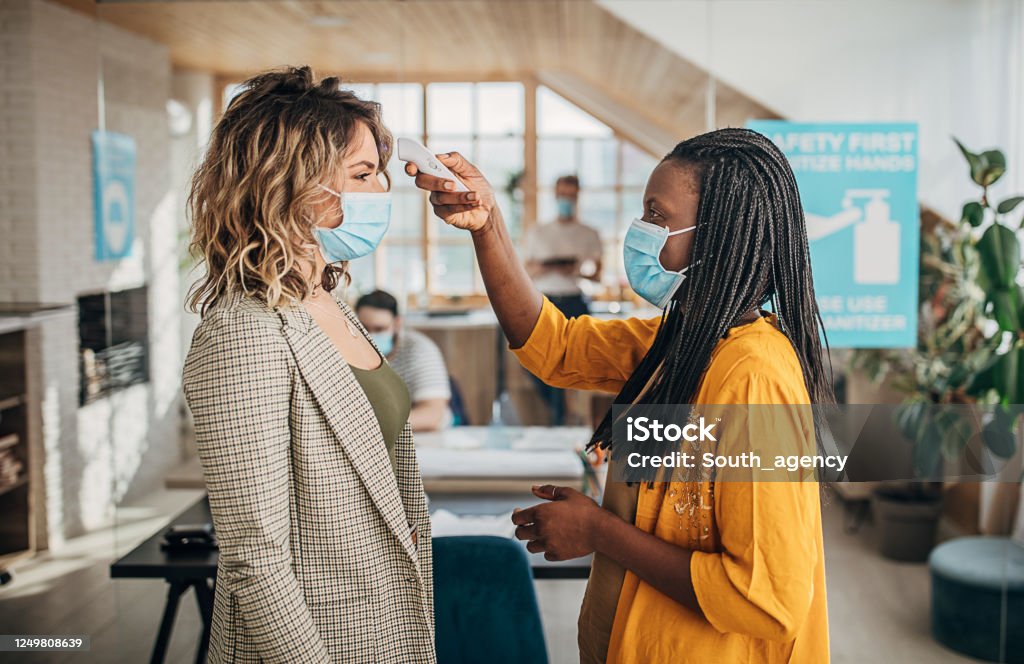 Two women using infrared thermometer for measuring temperature before entrance in office Colleagues with protective face masks using infrared thermometer for measuring temperature before entrance in office. Scanning for coronavirus or covid-19 symptom. Coronavirus Stock Photo